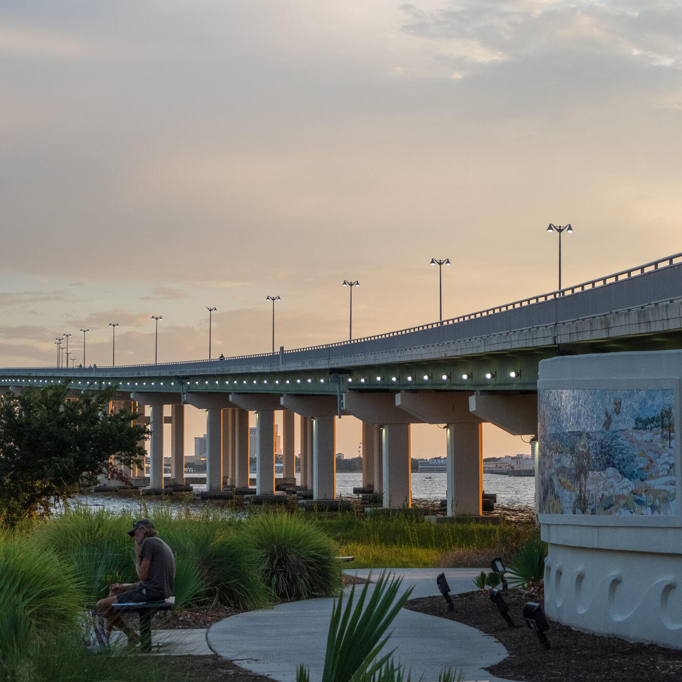 A large bridge at sunset. In the foreground, a man sits on a bench. To the right is a nautical mosaic mural.