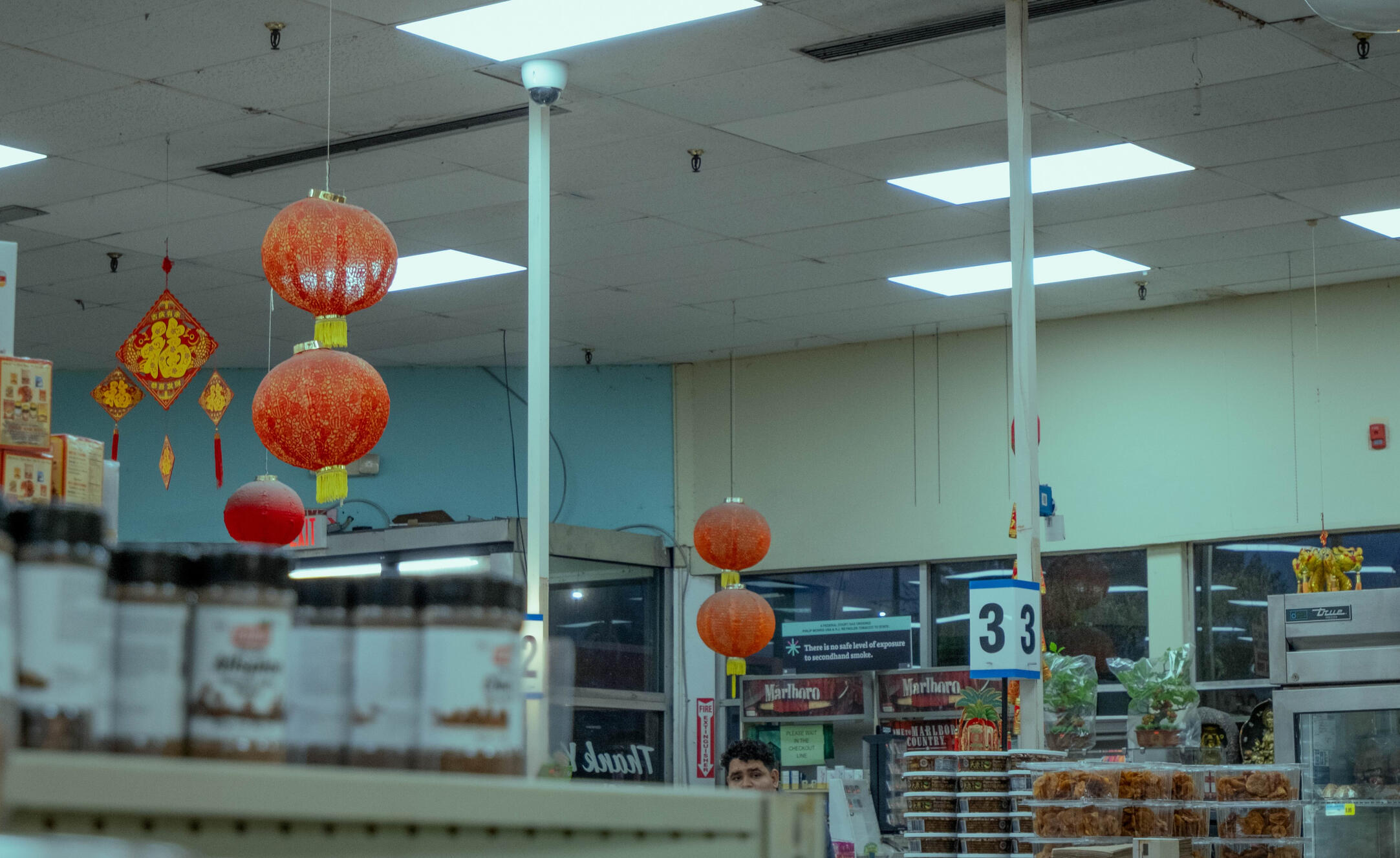A grocery store with fluorescent lighting and Chinese red lanterns hanging from the ceiling. At the bottom, a worker's face peers from behind a shelf of canned goods.