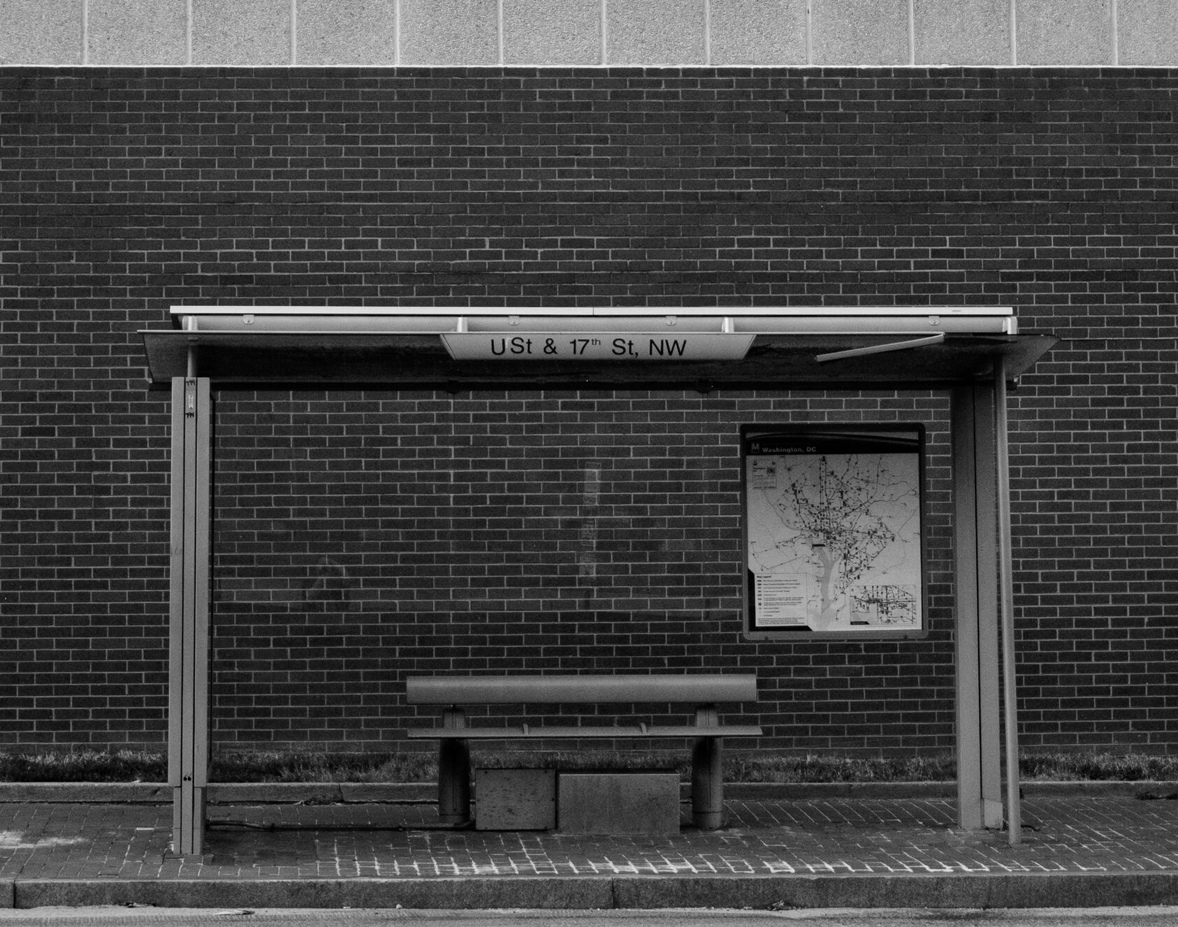Black and white photo of a covered bus stop with a metal bench. To the right is a city map of DC. The street marker at the top of the bus stop reads "U St & 17th ST, NW"