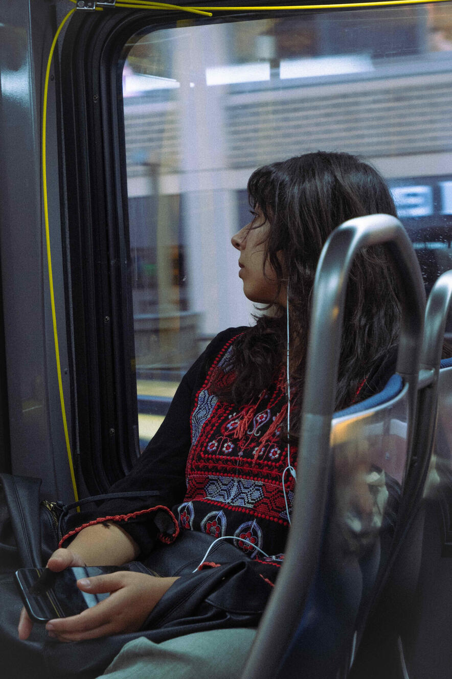 A young woman sitting on a city bus, wearing a traditional black Palestinian dress with white and red embroidery. She has one earbud in and the other hangs down. She is looking out of the window to the right.