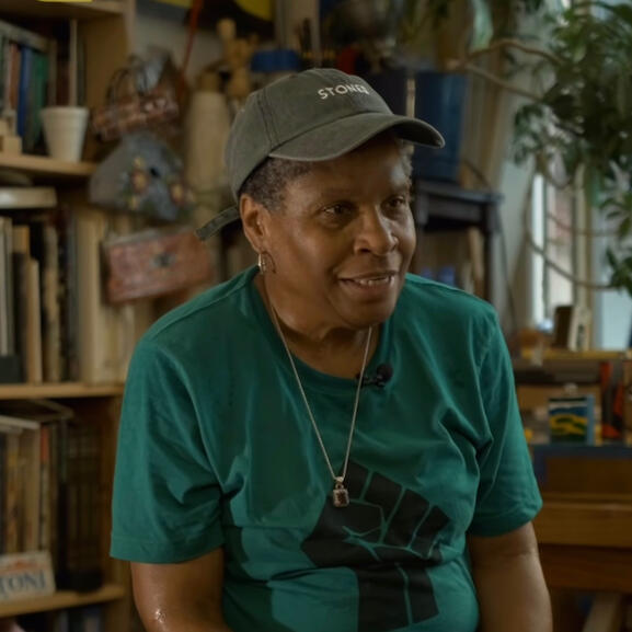 A woman in a baseball cap and green t-shirt with the power fist on it. She is sitting in an artist's studio with many books, paintings, and decor behind her.